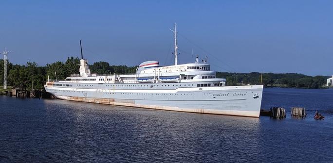 There is a large, steel lake-going ship docked on a blue lake, near a wooded shoreline