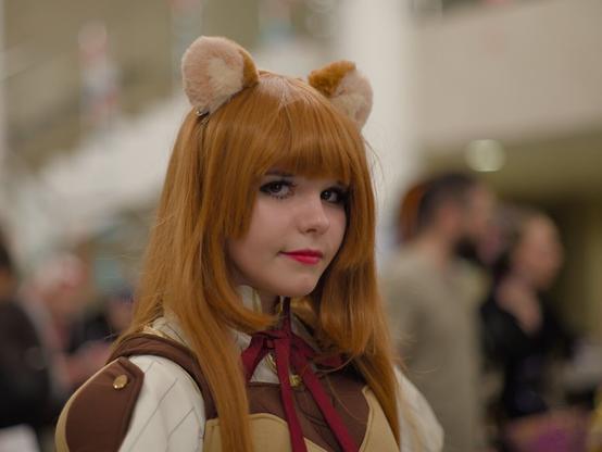 Head and shoulders portrait of a girl wearing white dress and brown and red camisole stylized to medieval one with brown shoulder straps. She wears red-brown wig with raccoon ears. She looks at the camera and smiles slightly.