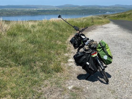 A very laden bike leaning on its kickstand  by the road side at the top of a descent towards water below