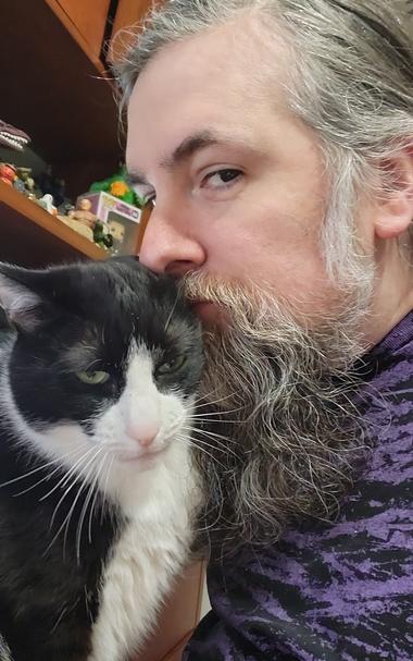 Me (gray hair, gray beard, purple shirt) next to my tuxedo cat-son Cubone, thinking about biting his ear gently because it's like furry taffy