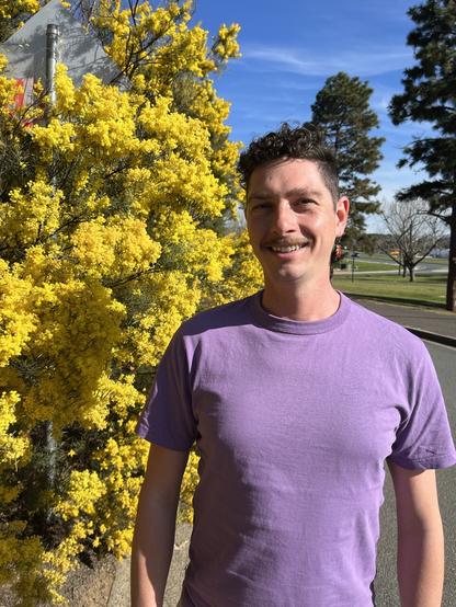 Ben standing outside wearing a lavender-purple teeshirt in front of a stunning yellow blooming wattle tree