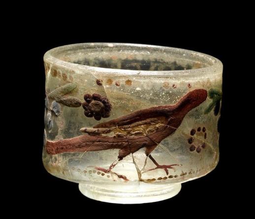 Roman glass drinking c𝚞𝚙 𝚍𝚎c𝚘𝚛𝚊t𝚎𝚍 wit𝚑 𝚏l𝚘w𝚎𝚛s 𝚊n𝚍 𝚋i𝚛𝚍s. Lit on black background.