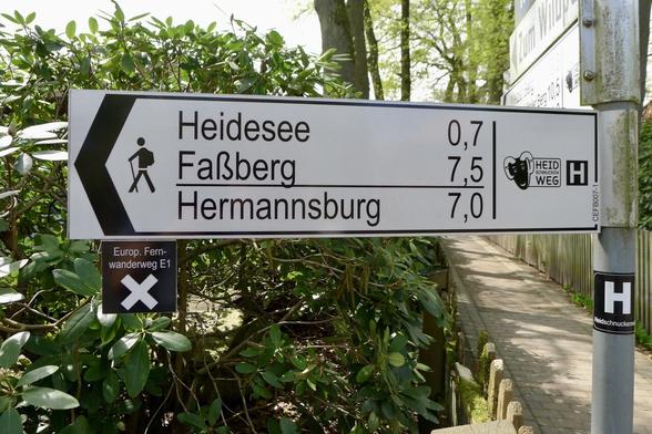 Ein Wegweiser mit mehreren Orts- und Entfernungsangaben. Heidesee 0,7 - Faßberg 7,5 - Hermannsburg 7,0. Außerdem sind Wegmarkierungen des Europäischen Fernwanderweges E1 und des Heidschnuckenweges angebracht.