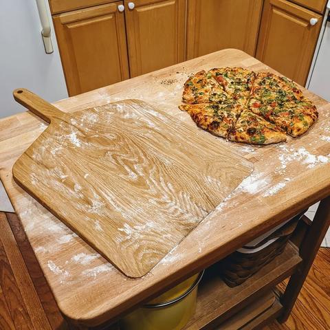 A white oak pizza paddle sits next to a cooked pizza on a kitchen island. Flour is everywhere.