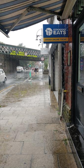 The photographer is standing under a shop awning out of the torrential rain. The road and pavement are glistening wet and a stream of water pours down the awning onto the wall.