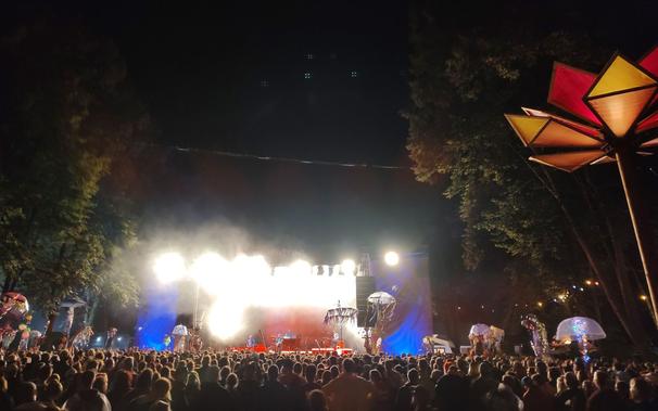 Große Bühne mit Lightshow und einer Band. Davor jede Menge Menschen. Einige davon halten Totems in die Höhe. Rechts und links Bäume.