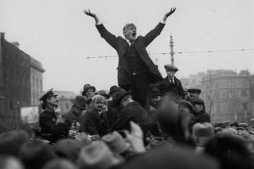 A photograph of James Larkin standing above a crowd with his arms aloft and his mouth open in mid-speech.