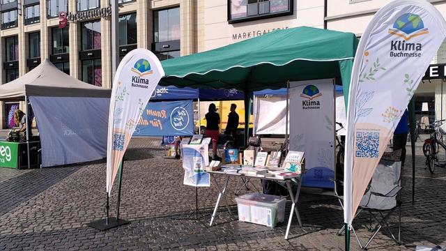 Foto: Stand der Klimabuchmesse (mit Beachflags und Rollup) steht unter einem grünen EasyUp-Zelt. Kopfsteinplaster darunter, im Hintergrund Geschäfte.