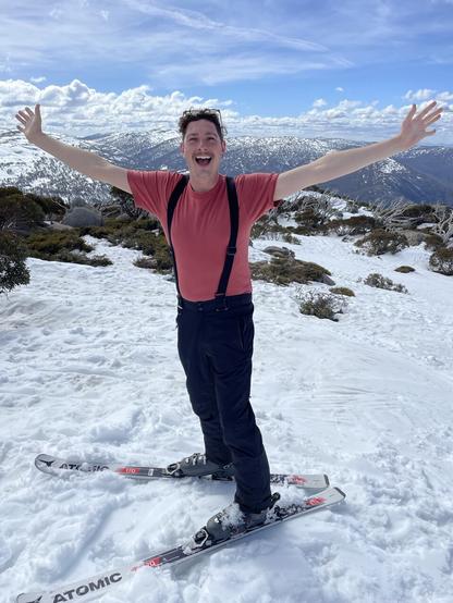Ben standing in the snow on skis, hands in the air, backdrop of snowy mountain ranges
