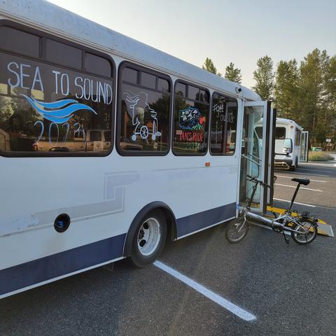 Photo of a white shuttle with wheelchair ramp extended and a small folding bike parked next to the ramp. On the windows, the words Sea to Sound 2023, Ian's Rde 2023, #GetOutside, and a picture of Ian on his wheelchair wearing his traditional checkered Vans on his feet with his distinctive dreadlocks flying out behind.