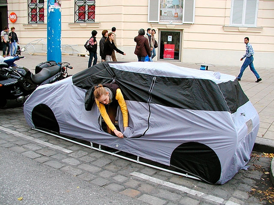 Exhibit by Michael Rakowitz in Vienna. A car-shaped tent has been constructed by the roadside, occupying a parking space. Someone in a yellow sleeved jacket is leaning out, fastening a zipper. In the background, pedestrians are walking on the sidewalk.