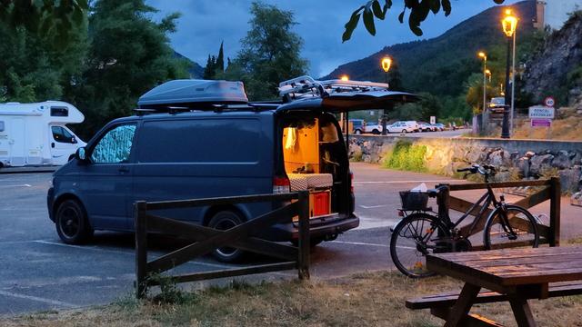 La furgo, aparcada en un pàrquing amb terra esfaltat i tot ben acabat, amb la bicicleta descarregada contra un barana però res estès. Llum de quasi nit amb fanals, es veu llum dins la a
furgo, de perfil i amb el portó obert.