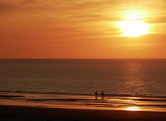 Sonnenuntergang am Meer, das Meer wirkt glatt. Oranger Himmel, der sich im nassen Sand spiegelt. 2 Reiter an der Wasserlinie.