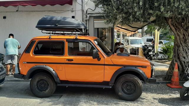 Side view of orange Lada Niva 4x4 parked on street by olive tree