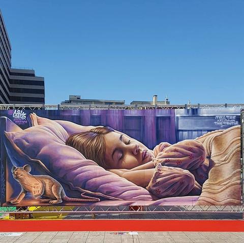 Streetartwall. On a long exterior wall on a metal fence, is sprayed / painted mural of a sleeping child. The brown-haired girl lies with closed eyes on a purple pillow. She has put her hands under her head. She is half covered with a beige blanket. On the far left of the picture runs a small brown mouse with a luminous pendant around the neck.