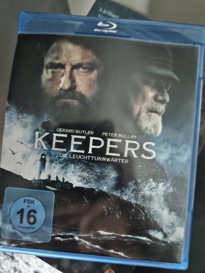 Bluray-Hülle zum Film Das Cover ist Standard. Die beiden Hauptrollen im Zentrum thronen über einer schattenriss-artigen Szene, die eineb Leuchtturm in tosender Brandung zeigt.