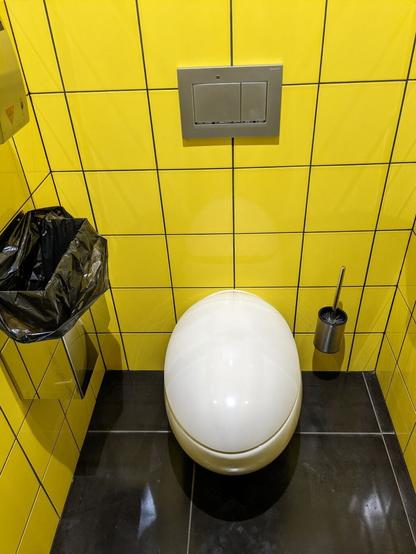 Eine geschlossene Toilette in Form eines Eis vor gelben Wandfliesen. Die Bodenfliesen sind schwarz. Auch zu sehen (alles silbern): Klobürste, Mülleimer und Spülung.