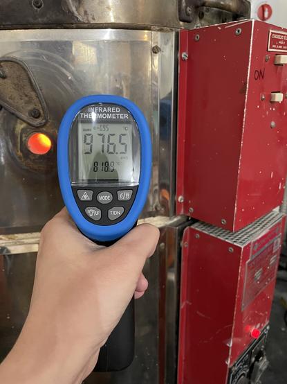 An infrared thermometer reading 976.5 degrees Celsius with a kiln in the background. One of the kiln peeps is open and you can see the bright orange of the kiln atmosphere through it