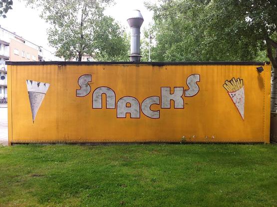 A long rectangular yellow cabin outside with the word SNACKS on it. There is also a drawing of an ice cream cone on  one side and some fries on the other. The cabin is located on a patch of green grass. On top of the cabin is an aluminium chimney.