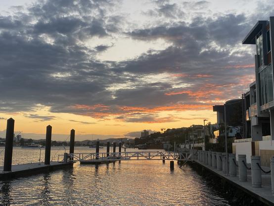 Brisbane river at sunset. Grey scattered clouds above, with the undersides of the further ones tinged with orange. The horizon is yellow. To the right are a walkway and riverside apartments. Lapping water can be heard (although I have headphones on).