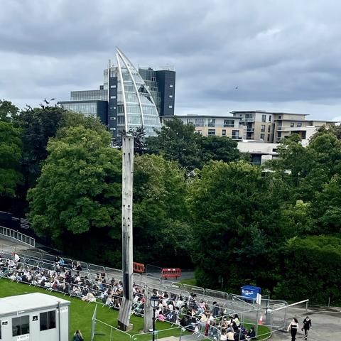 Long distance shot from above: a milky grey sky. Modern buildings with glass and varied facades on the horizon. A wall of full-leafed trees in the mid-ground. A tall abstract wood sculpture. In front, on a lawn corralled between lanes of temporary fencing, a hundred or so young people line-up waiting on a concert to start.