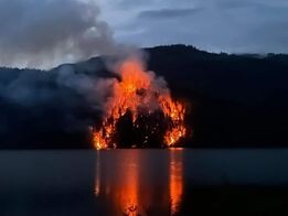 A photo of a wildfire across Lake Whatcom from the morning of August 29, 2023. The wildfire is noticeably bigger than in the previous photo, showing a clear ring of fire surrounding a dark burned-out area in the middle. Photo by Bradley James Lockhart