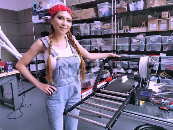 Naomi Wu, a well known maker originally from China, in her Workshop, wearing blue jean Dungarees, whilst showcasing the "Printmill", A 3D Printer that has a converyor belt and thus can churn out either multiple pieces in one go without human intervention or parts that are theoretically infinitely long in one axis [horizontal]...