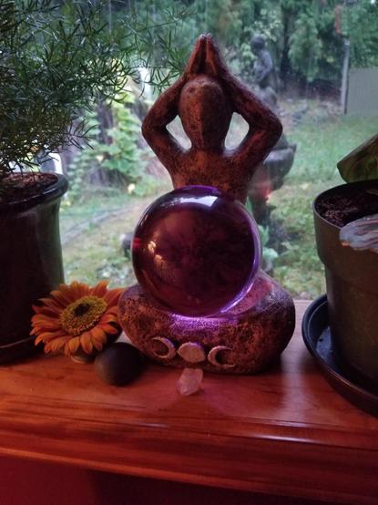 A Goddess statue with a triple moon sign on the bottom sits in a window with a lavender coloured glass ball on her lap. A sunflower is beside, a small quartz crystal and a smooth black rock in front. A lacy potted fern sits beside her. Through the misty window, a trailing pumpkin vine can be seen.