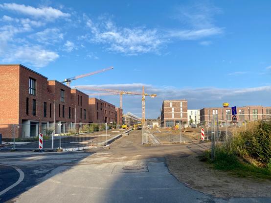 Zwischen rot geklinkerten  Wohngebäuden sieht man, wie die Zufahrt vom alten Dorf am Entstehen ist. Im Hintergrund Kräne.