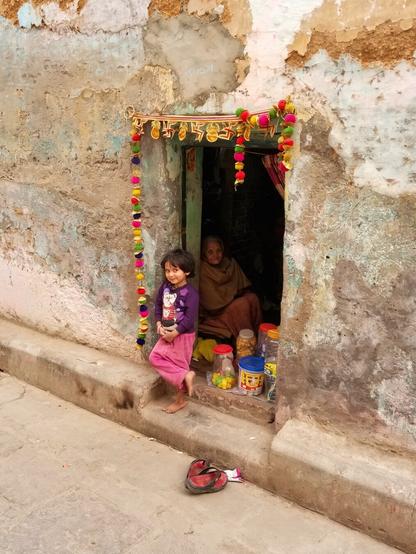 A family of candy sellers in the alleys of Varanasi. A small girl stands in a doorway with her grandmother barely visible inside.