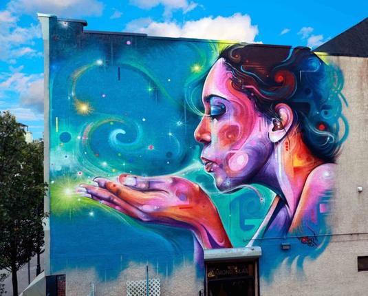 Streetartwall. On the outer wall of a modern building is sprayed/painted a colorful, wild mural in pop art style. From the side you can see a young dark-haired woman blowing over her hands and forming a heart with the wind. The background is designed in bright blue with yellow star sparkles. The young woman is also dipped in purple, pink and turquoise color veils. A fabulously beautiful mural.