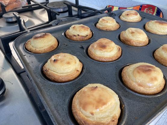 A cupcake tray on a hob, filled with freshly baked pasteis de nata, each a browned and crispy puff pastry case filled with a risen, pale yellow custard with some caramelisation on top.
