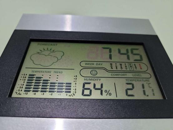 A digital clock/thermometer, liquid crystal screen with black data on a greenish-gray background and black border, mounted on a green wall, depicting 21.1 degrees Celsius, 64% humidity, time