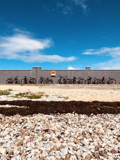 Lots of bike against a grey wall, blue sky, sand and stones in the foreground