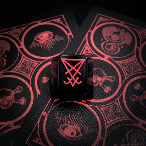 photograph of an artisan keycap displaying the sigil of Lucifer, the cap sits on top of two playing cards showing symbols of secretive societies