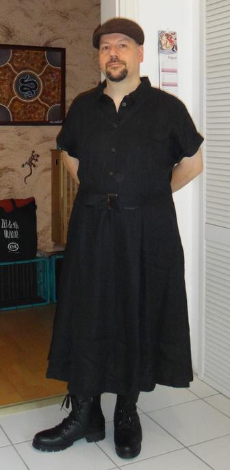Foto von mir, einem leicht untersetzten Mann mittleren Alters mit dunklen Augen und dunklem Kinnbart. Trage ein langes schwarzes Leinenkleid mit geknöpftem, hemdartigem Oberteil und weitem Rock. Außerdem eine dunkelbraune Filzmütze und schwarze Schnürstiefel.