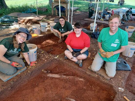 Four people sit on the edges of two archaeological units, all are smiling at the camera. All people hold a small trowel in their hands. There is sunlight on the grass behind them, and another group of people digging in other units in the distance.