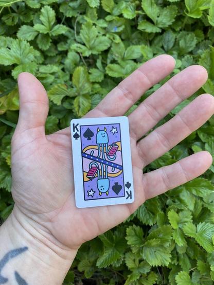 A tiny playing card in my outstretched hand, it is smaller than my palm. Shows the King of spades who is a blue, smiley, horned squid sort of creature reading a book.