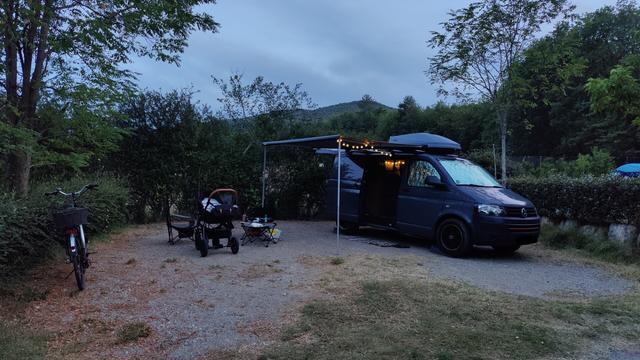 La furgoneta viatgera aparcada en una parcel·la amb terra de grava i uns arbustos baixos separadors. Es veuen pocs arbres. El toldo, cadires, taula, cotxet i bicicleta estan estesos, i del toldo penja una cadena de llums ornamentals. És vespre, la foto és fosca.