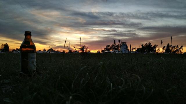 Sunset at airfield. Planes and tower in the background, bottle of beer in front. Grass growing.