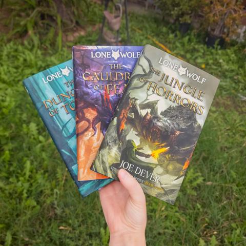 Photograph of Lety's hand holding up three books in her garden. The books are "The Jungle of Horrors," "The Cauldron of Fear," and "The Dungeons of Torgar" from the Lone Wolf gamebook series by Joe Dever. These books are the hardback Definitive Editions printed by Holmgard Press and are very big, making Lety's hands look very presidential in comparison.