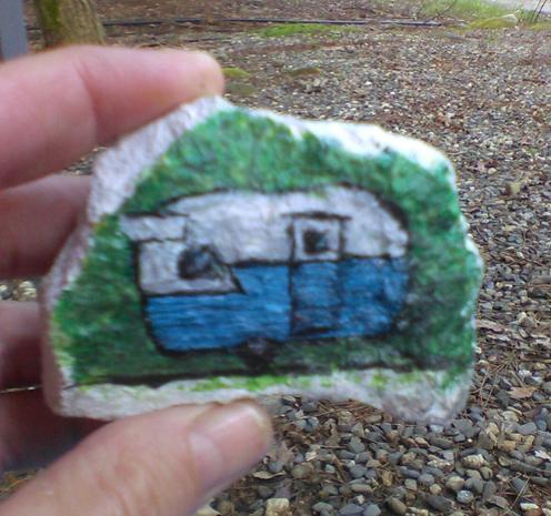 a small rock, painted white with a blue & white Shasta Canned Ham travel trailer on green background in acrylic paint.