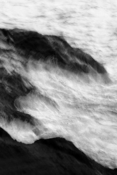 Black and white portrait format image using intentional camera movement and a slow shutter speed to blur waves crashing against dark rocks, displaying the power of the sea and the rhythm of the tides