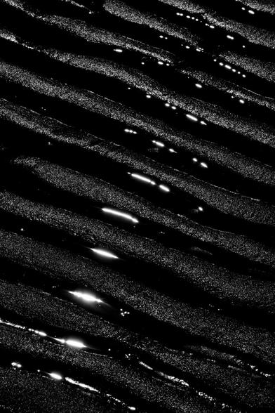 Black and white portrait format image showing parallel ripples in dark sand running diagonally across the frame, with sunlight glinting off the water left behind at low tide