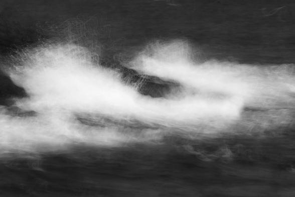 Black and white landscape format image using intentional camera movement and a slow shutter speed to blur waves crashing against dark rocks, displaying the power of the sea and the rhythm of the tides