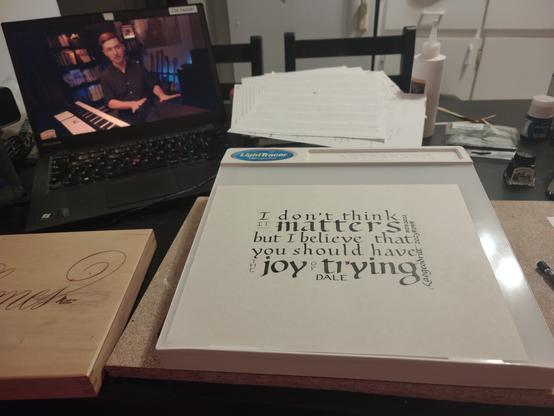 Photo of a messy artist's table. Foreground, a piece of gold-ish parchment paper. In calligraphy in the foundational hand and black ink, it reads, "I don't think it MATTERS, but I believe that you should have the JOY OF TRYING." Quote attributed to Dale. Signed Laogeodritt, September 2023, hashtag DB 2022. Various bits of art equipment around the piece, with a laptop screen in the background showing a video of music theory YouTuber Adam Neely.