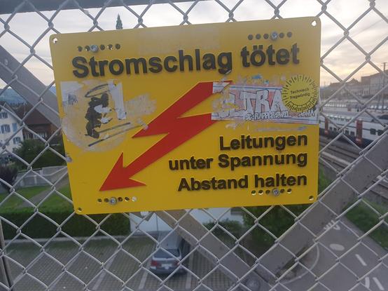 Ein gelbes Warnschild, "Stromschlag tötet - Leitungen unter Spannung - Abstand halten", auf dem einige Stickerreste kleben.
Außerdem ist da ein gelber Inspektionssticker: "technisch fragwürdig. Bei diesem ist kein letztes/nächstes Prüfdatum indiziert.
