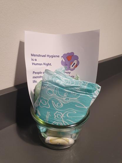 Ein Einmachglas, das einige Binden und Tampons sowie einen Informationszettel beinhaltet, steht auf einer grauen Ablagefläche.