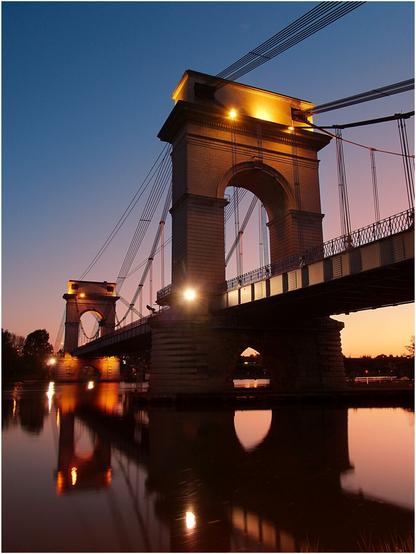 Photo de nuit, au crépuscule, du pont du Port-à-l'Anglais. Sur la photo, prise depuis une rive, on voit, en contreplongée, les deux pylônes de pierre, respectivement en premier plan et en arrière-plan. La luminosité ambiante étant faible, la pose est longue. Cela a pour effet de figer l'eau du fleuve. Le reflet du pont apparait comme dans un miroir