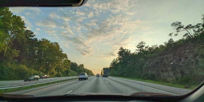 Photo of a sunset over a highway surrounded by trees on the left and a rocky hill on the right.